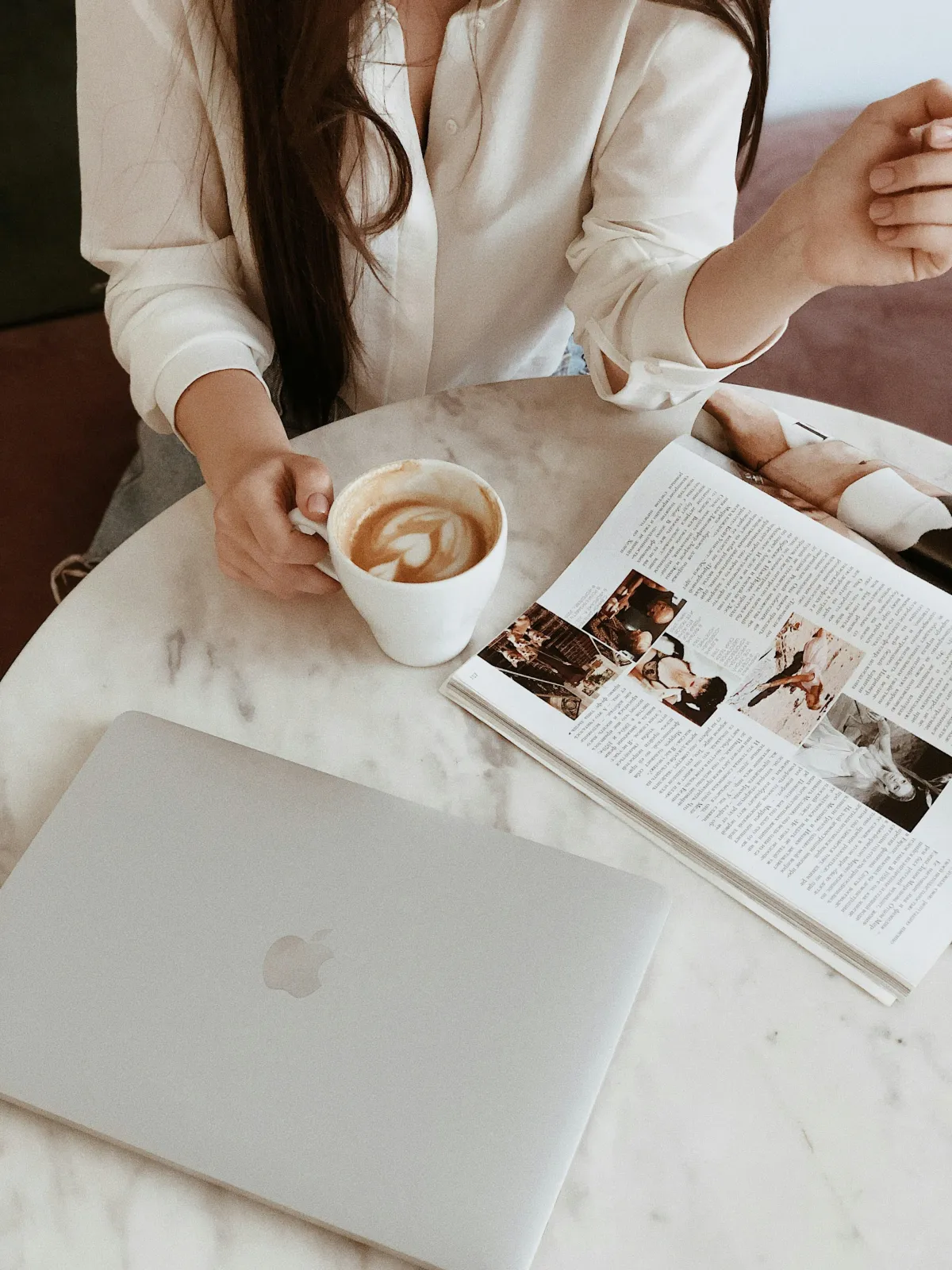 Overhead shot of a woman enjoying coffee at a marble table with a closed laptop and magazine, reflecting intentional living and productivity.