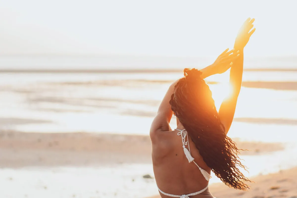 Back view of a woman on a beach at sunset, raising her arms in relaxation, symbolizing freedom, mindfulness, and connection with nature.