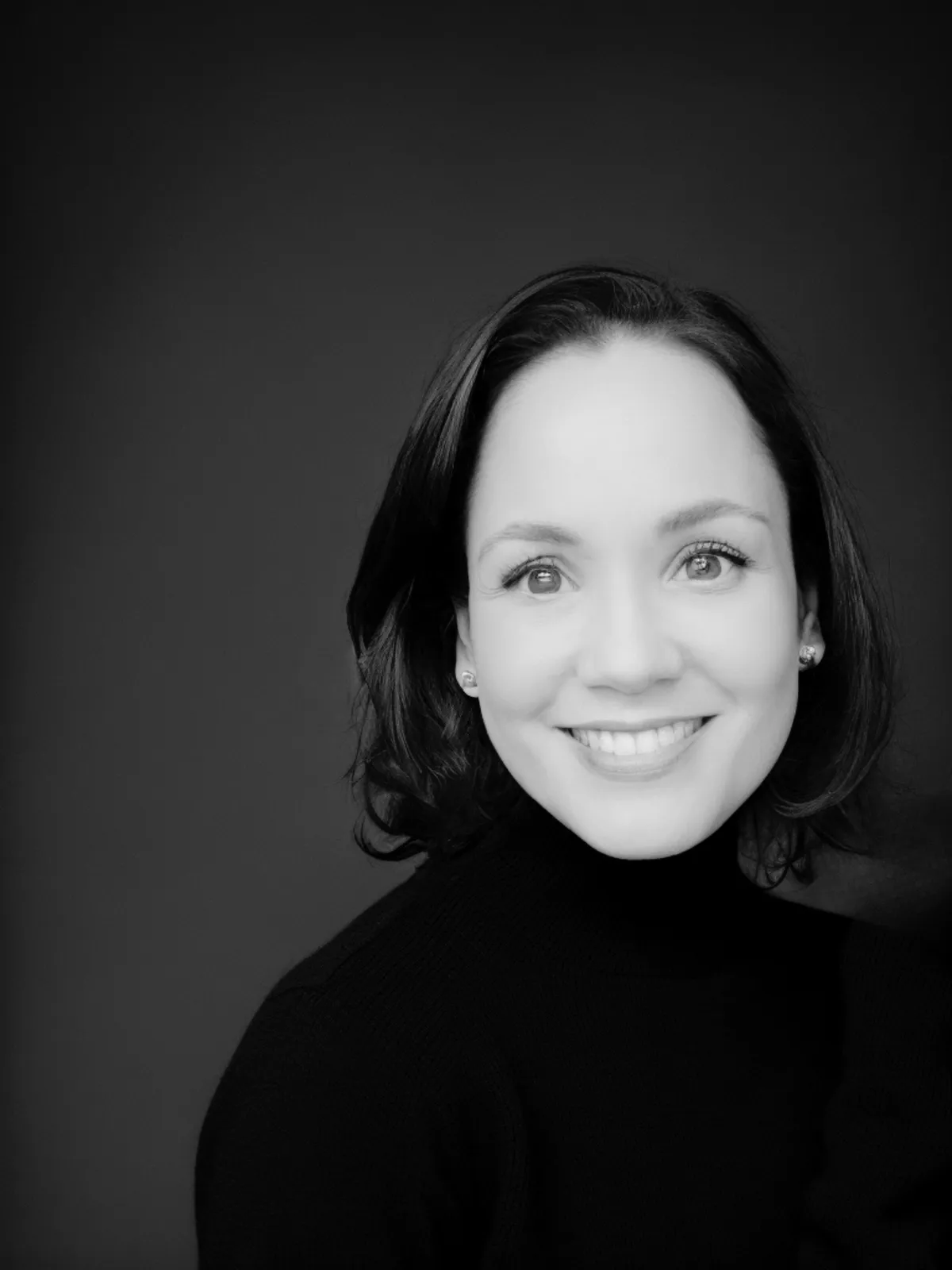 Black and white portrait of Jeannette, the founder of Now She’s Living, smiling warmly in a black turtleneck, representing confidence, empowerment, and professionalism.