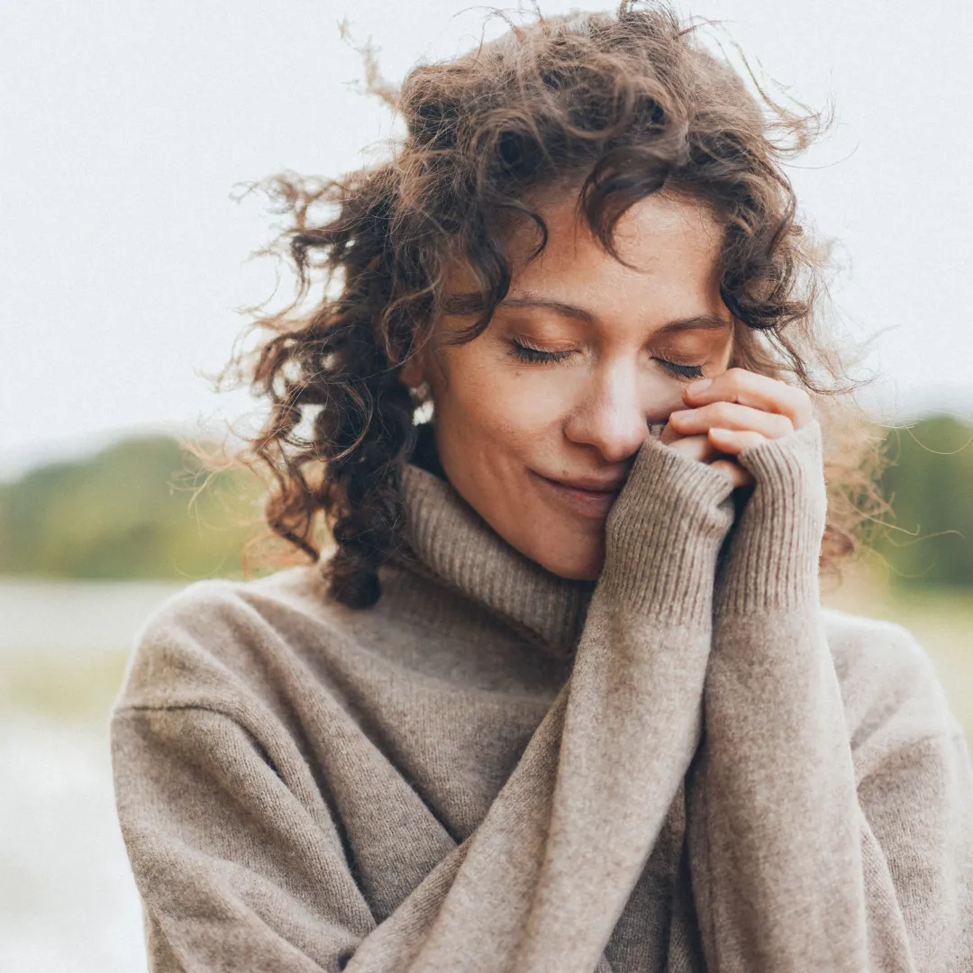 A serene woman with curly hair smiles gently with her eyes closed, wrapped in a cozy beige sweater, embodying a sense of peace, self-love, and quiet contentment.