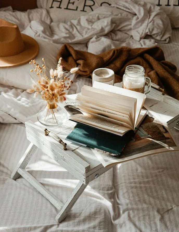 A cozy bed setup with a white tray table holding an open book, a jar of coffee, a candle, and dried flowers in a vase, with a beige hat and soft blankets in the background, evoking warmth and relaxation.