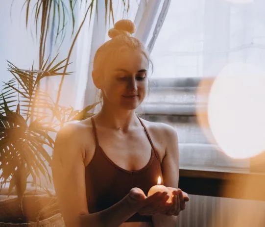 A woman seated in a serene room holding a lit candle, with soft natural light surrounding her. She appears calm and focused, embodying mindfulness and spiritual connection.