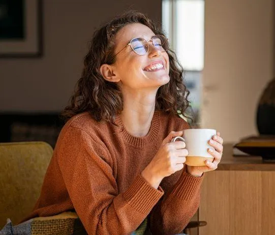 A smiling woman in a cozy, neutral-toned sweater, sitting in a relaxed posture while holding a coffee cup. She exudes a sense of peace and joy, symbolizing self-care and contentment.