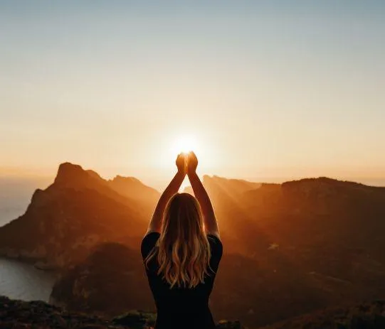 A woman standing on a mountain at sunset, holding her arms up to the sun. The golden light surrounds her, representing inner peace, empowerment, and a connection to nature and spirituality.