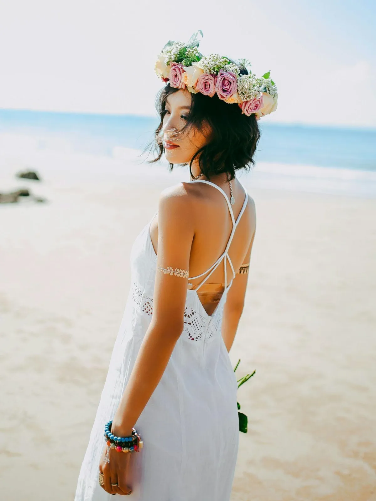 Woman wearing a white dress and floral crown, standing on a beach with a serene expression. Ideal for themes of femininity, peace, and natural beauty.