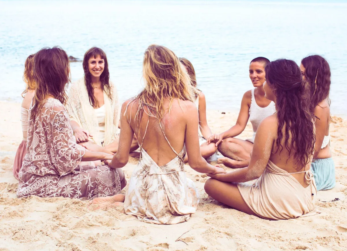 Two women smiling and wrapped in a colorful blanket on a sunny beach, enjoying the breeze and each other's company.