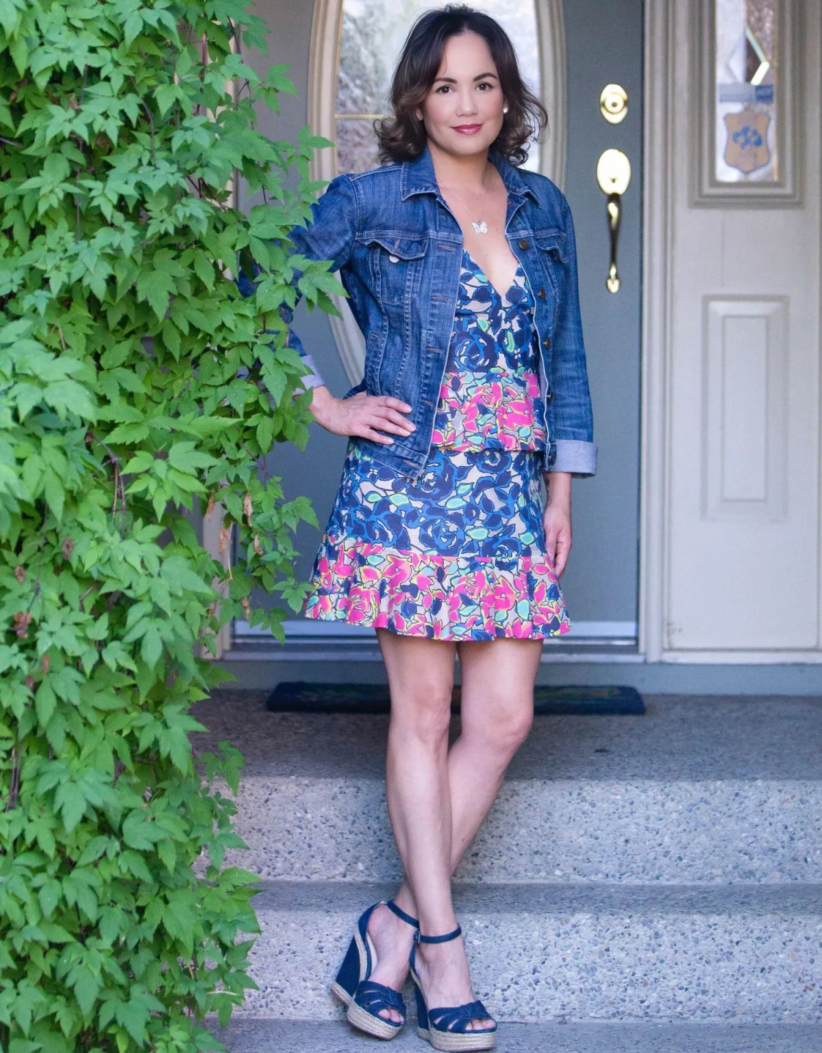 Jeannette, founder of Now She’s Living, standing confidently in front of a doorway. She is wearing a vibrant floral dress, a denim jacket, and navy wedges, surrounded by greenery, exuding a warm and approachable energy.