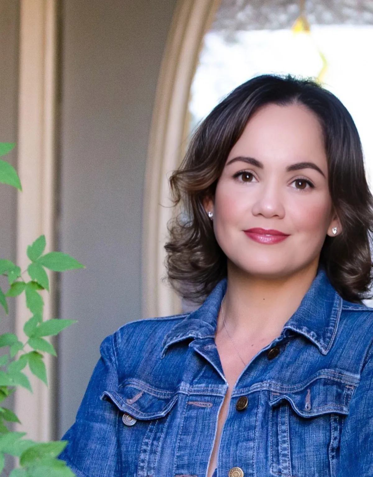 Jeannette, the founder of Now She’s Living, smiling warmly with a calm and welcoming presence. She is wearing a denim jacket and standing near a softly lit arched window surrounded by greenery.