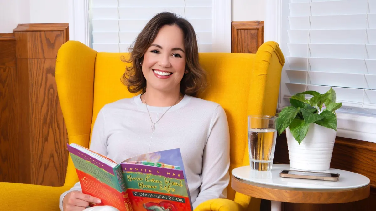 Jeannette Hounjet, founder of Now She’s Living, sitting in a vibrant yellow chair with a warm smile, holding Louise Hay's You Can Heal Your Life companion book. A peaceful and inviting scene, symbolizing healing, self-discovery, and spiritual growth.