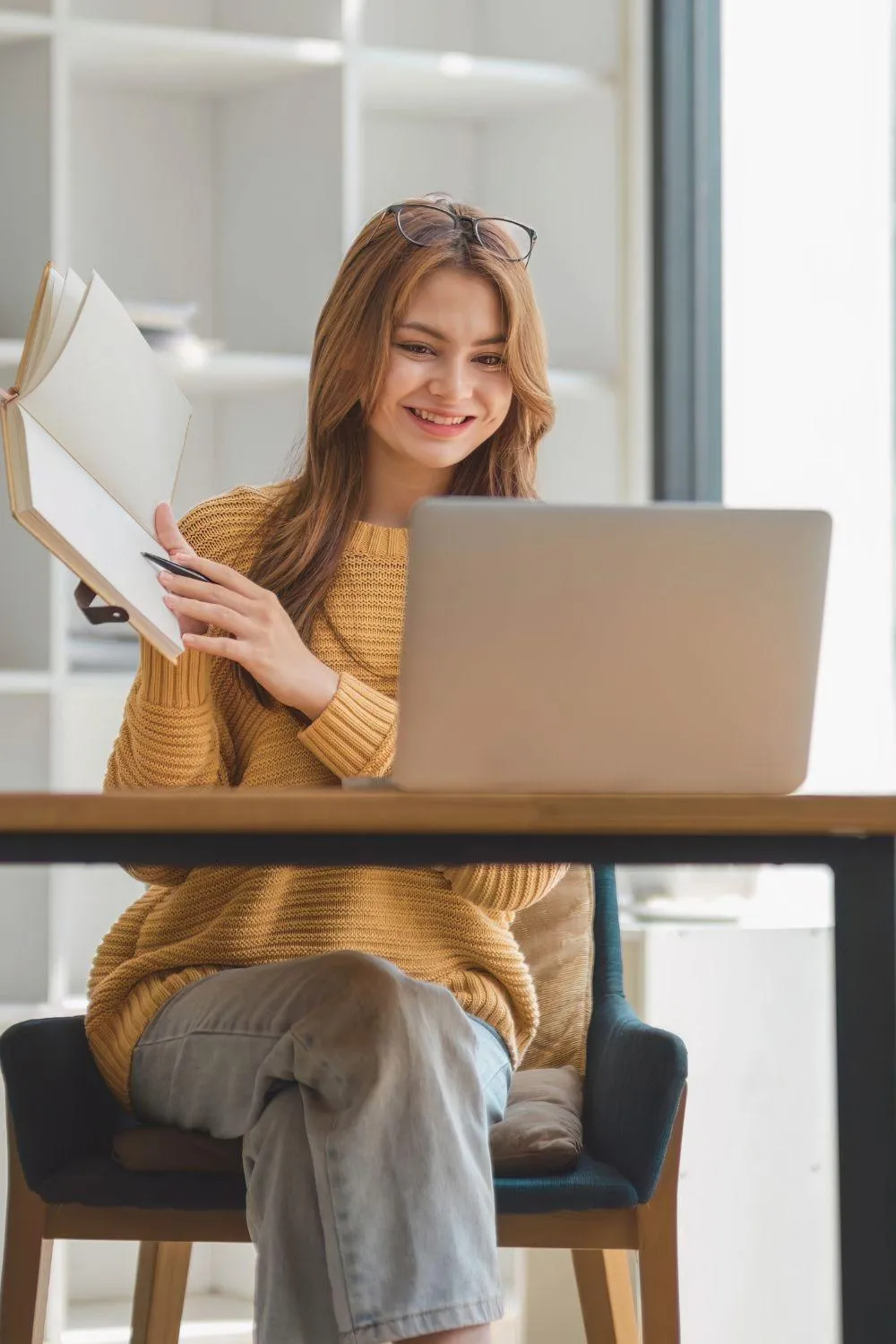 A smiling woman in a cozy yellow sweater participates in an online workshop from her home, holding a notebook while engaging with her laptop. The bright, inviting space reflects connection, learning, and personal growth