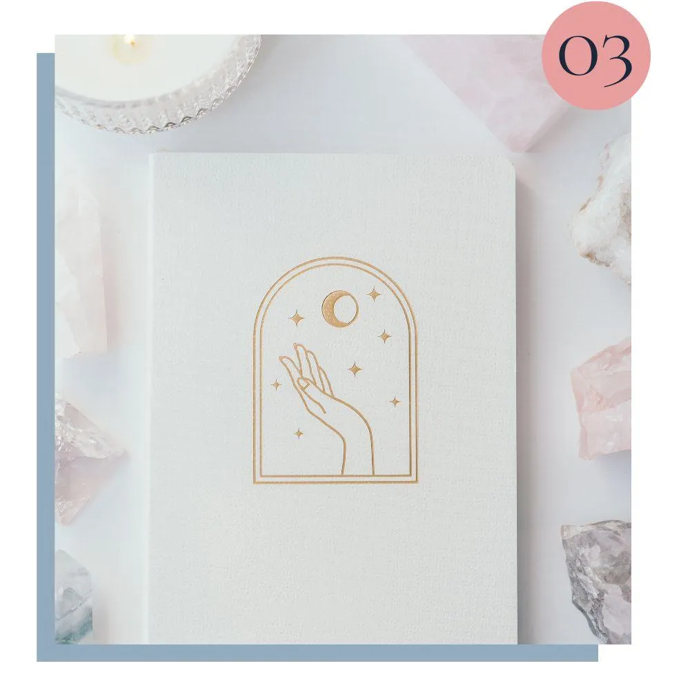 A minimalistic journal with a golden hand, crescent moon, and stars design, surrounded by crystals and a candle, symbolizing reflection, growth, and sacred space.