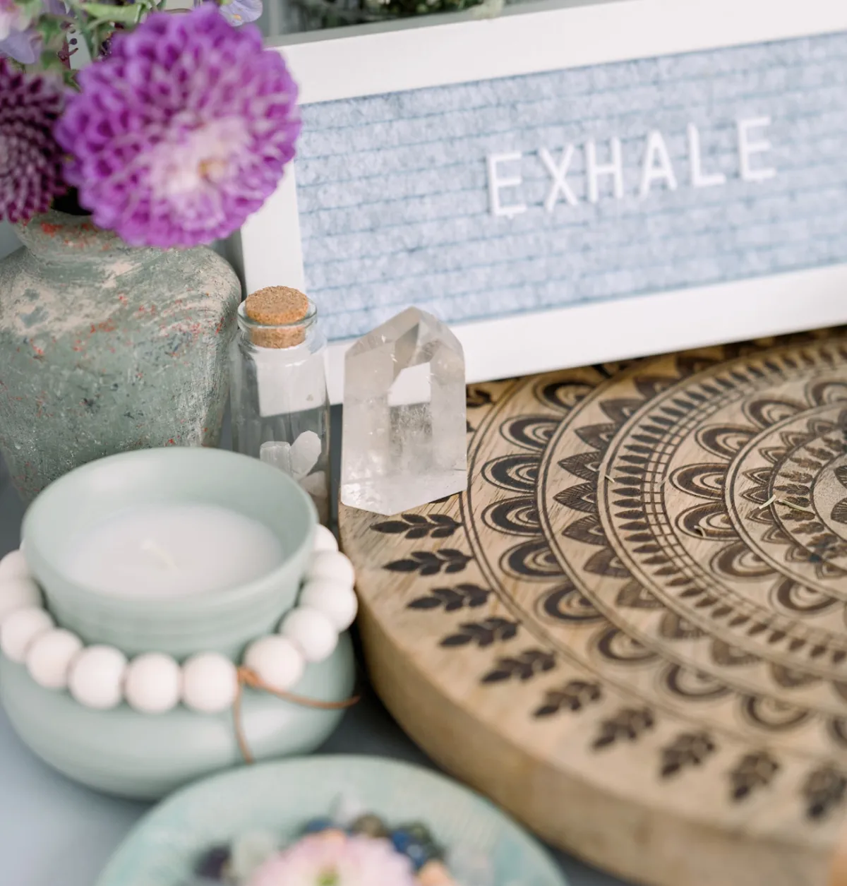 A serene arrangement featuring a candle, crystals, flowers, and a decorative mandala, with a sign reading 'Exhale,' symbolizing relaxation, healing, and intention setting.