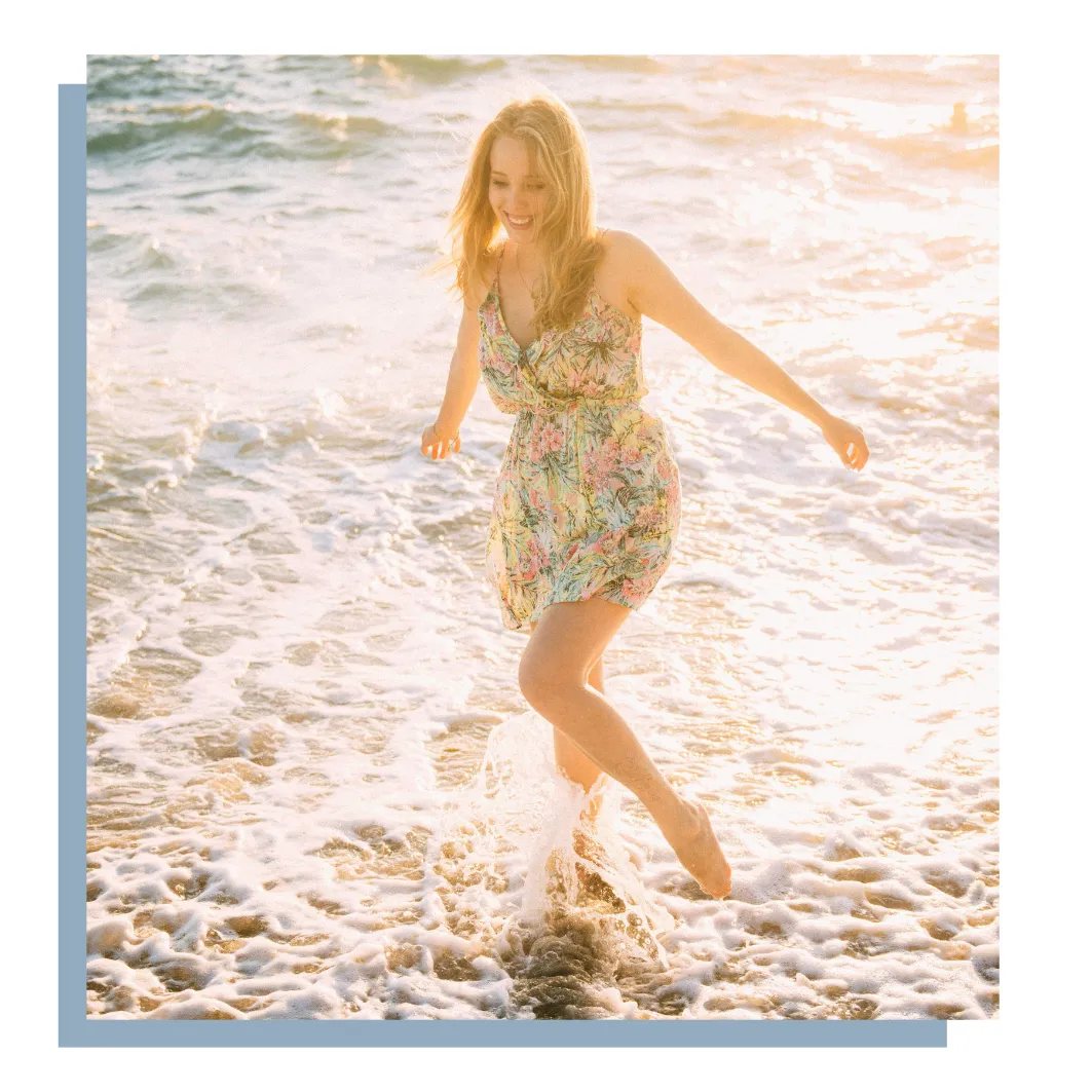 A joyful woman in a floral dress walking through ocean waves at sunset, symbolizing freedom, self-love, and transformation