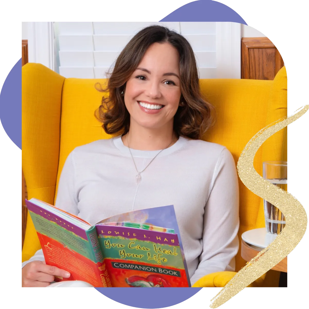 Jeannette, a self-love coach for sober women, smiling warmly while sitting in a bright yellow chair and holding the Louise Hay book You Can Heal Your Life® Companion Book.