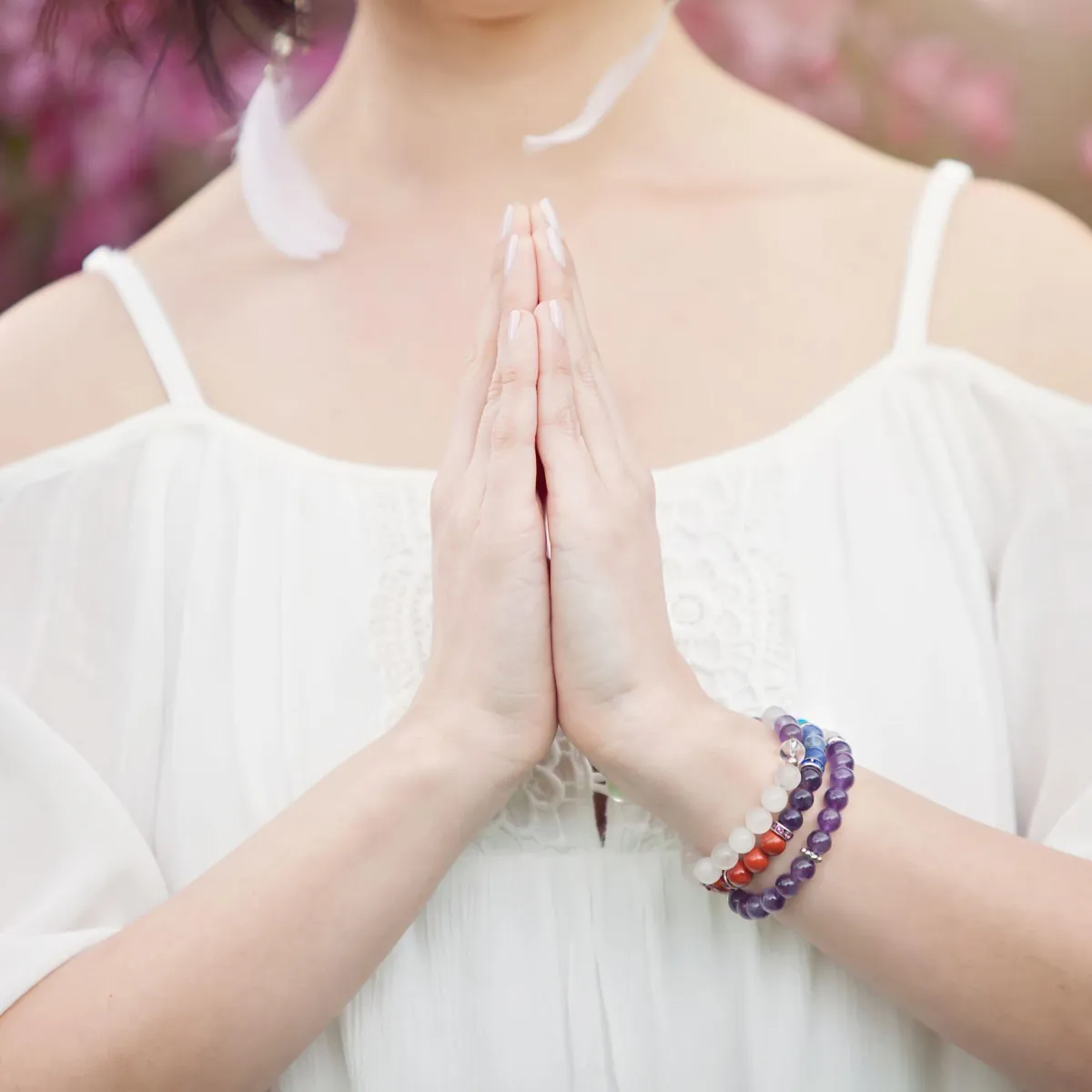 Close-up of a woman’s hands in prayer pose, wearing colorful gemstone bracelets, with soft light and a backdrop of pink flowers, evoking peace and spiritual focus.