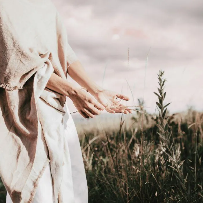 A person wearing light-colored clothing stands in a field of tall grass, with hands outstretched as if feeling or interacting with the plants, suggesting connection and flow.