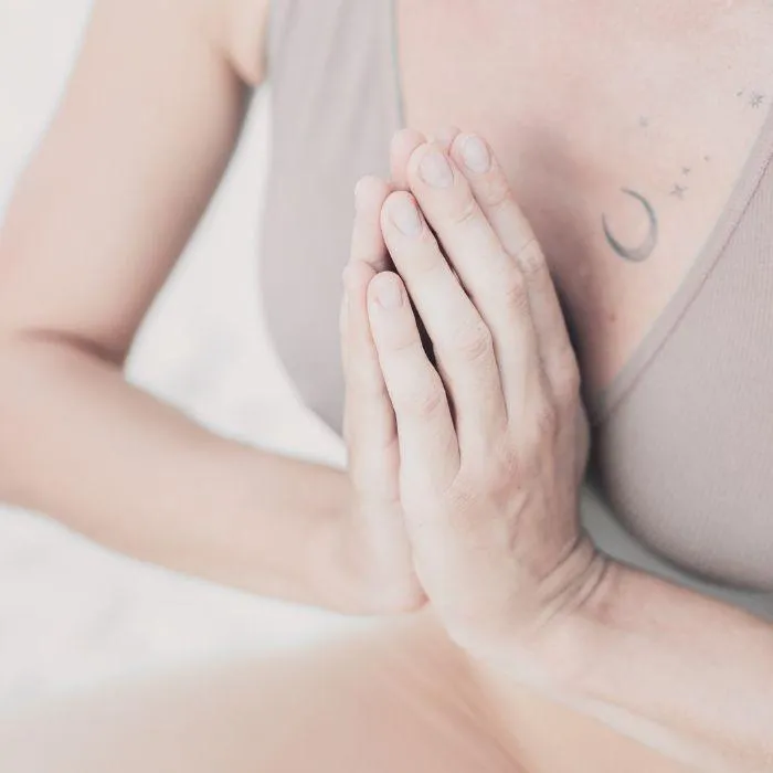 A close-up of a person's hands pressed together in a prayer or anjali mudra position, resting at chest level, with a blurred soft background.