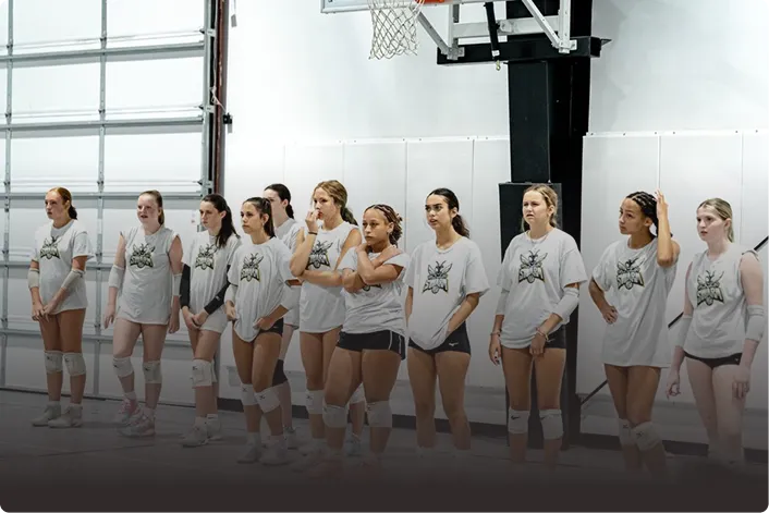 A girls' volleyball team in white and black uniforms gathered around their coach in a gym.