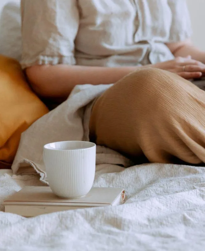 Woman sitting on a bed with a tablet, next to a book and coffee, symbolizing a relaxed approach to simplifying business tech solutions.
