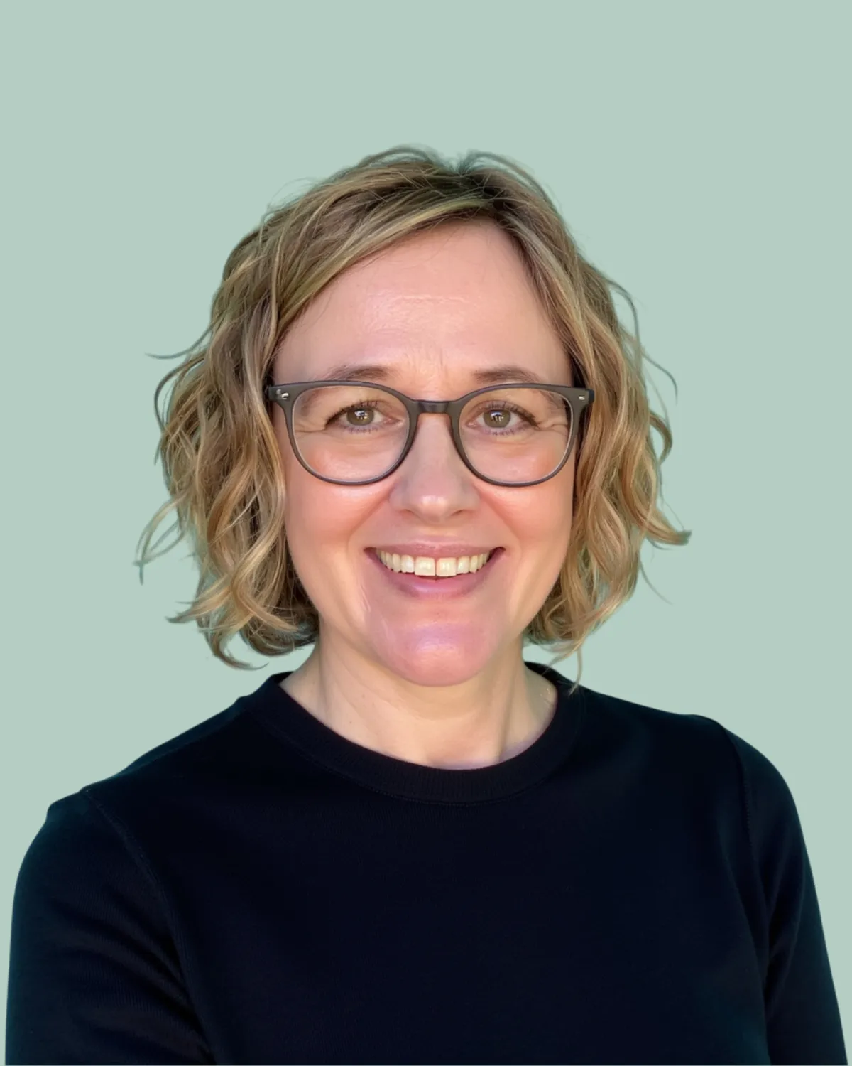 Picture of Emily Wilkinson, a woman with wavy hair wearing glasses and a black sweater. Mint green background.