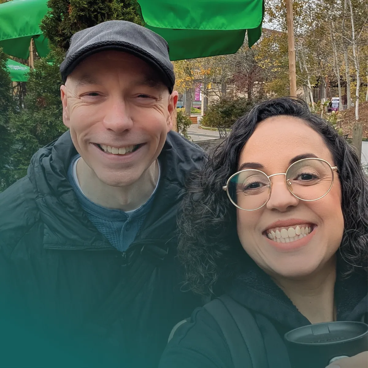 A bright, outdoor selfie of Rosicler and John Edward McGraw smiling warmly at the camera. John is on the left wearing a dark cap and jacket, while Rosicler is on the right wearing round glasses and holding a cup. They are standing near green patio umbrellas with autumn trees visible in the background. A teal color gradient overlays the bottom left corner of the image.
