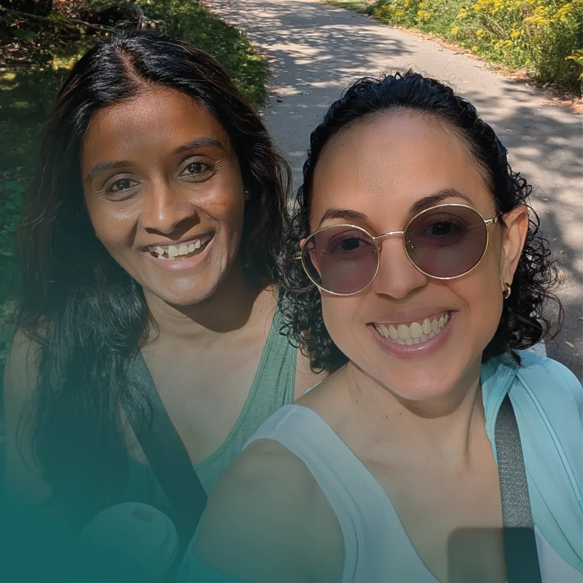 A bright, outdoor selfie of Rosicler and Prayalini (Pri) Sathanantham smiling warmly at the camera. They are standing together on a sunny, tree-lined walking path. Prayalini is on the left in a casual green top, and Rosicler is on the right wearing round sunglasses and a light top. A teal color gradient overlays the bottom left corner of the image.