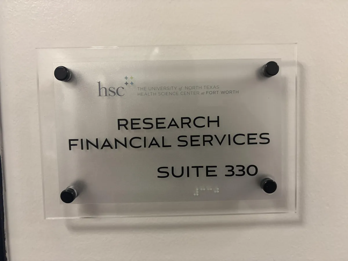 Clear acrylic office sign mounted on a white wall displaying ‘Research Financial Services Suite 330’ with the University of North Texas Health Science Center at Fort Worth logo