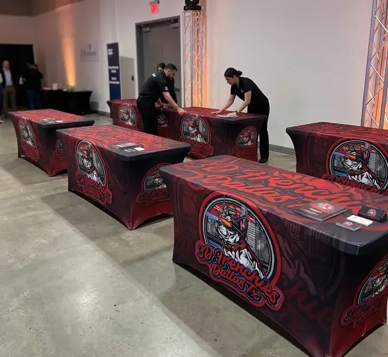 Two professional installers arranging multiple red and black custom-branded stretch table covers for "3D Frenchies of Dallas" at an indoor event venue
