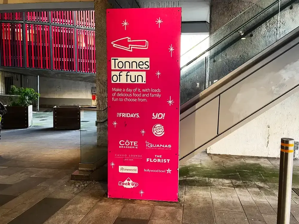 A bright pink vertical wayfinding sign for 'Tonnes of Fun' placed in an urban outdoor walkway to guide event attendees