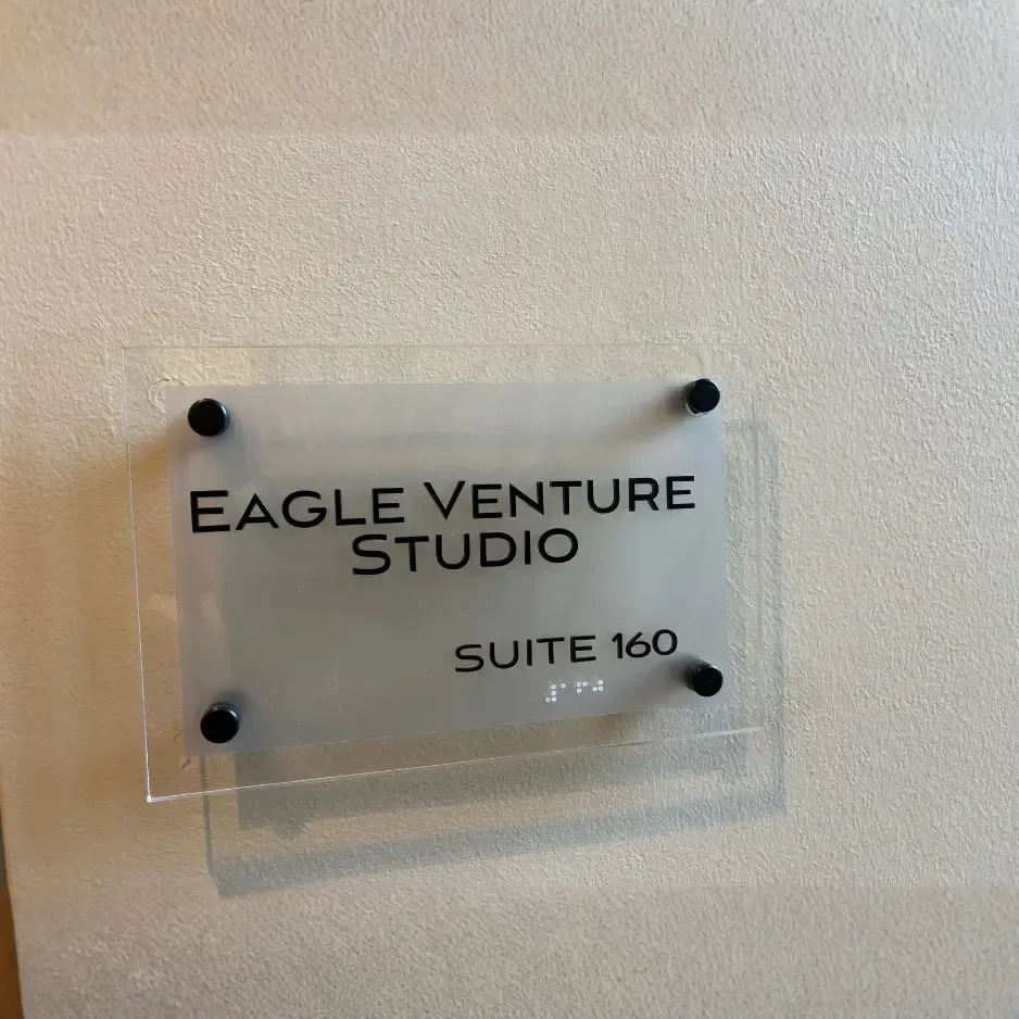 A professional clear acrylic suite sign for Eagle Venture Studio with silver mounting standoffs on a neutral office wall
