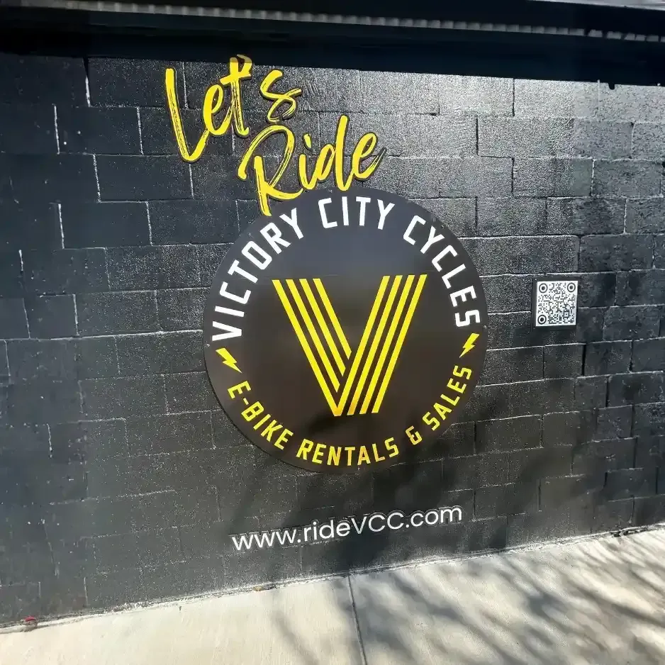 Bold yellow and white 'Let’s Ride' wall graphic for Victory City Cycles e-bike rentals on a black painted brick exterior
