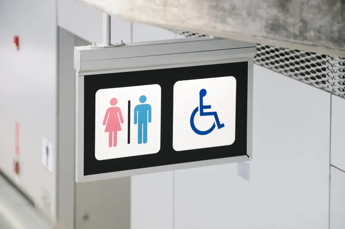 A double-sided projecting restroom sign featuring universal symbols for men, women, and accessibility, mounted in a clean corridor