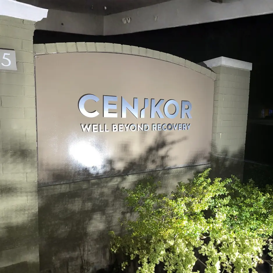Illuminated outdoor monument sign for Cenikor featuring silver lettering on a curved stone wall with evening landscaping