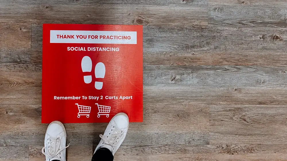 A red square floor decal on a wood-look surface with white text reading "THANK YOU FOR PRACTICING SOCIAL DISTANCING" and grocery cart icons