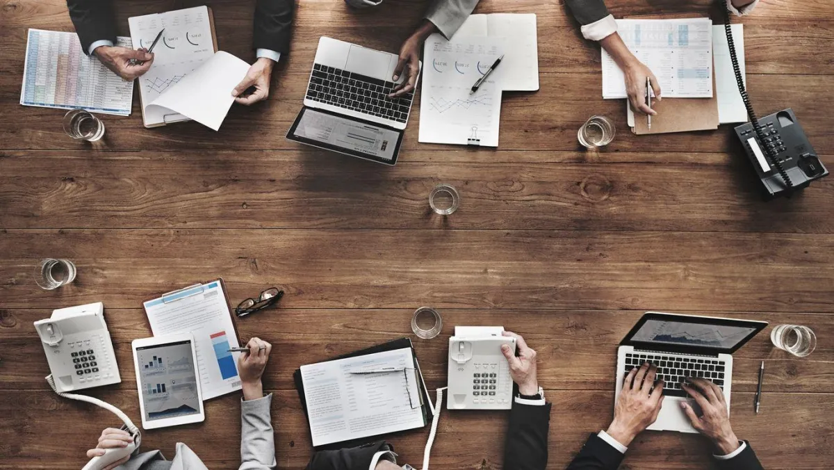Three diverse nonprofit leaders collaborating around a glass table with laptops and notes, bright natural light, modern office skyline visible, photorealistic style emphasizing teamwork and strategic planning.