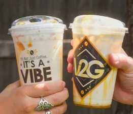 Two people clinking iced Vibes 2 Go white coffee drinks with caramel drizzle, showcasing V2G branding during a sunny outdoor moment.