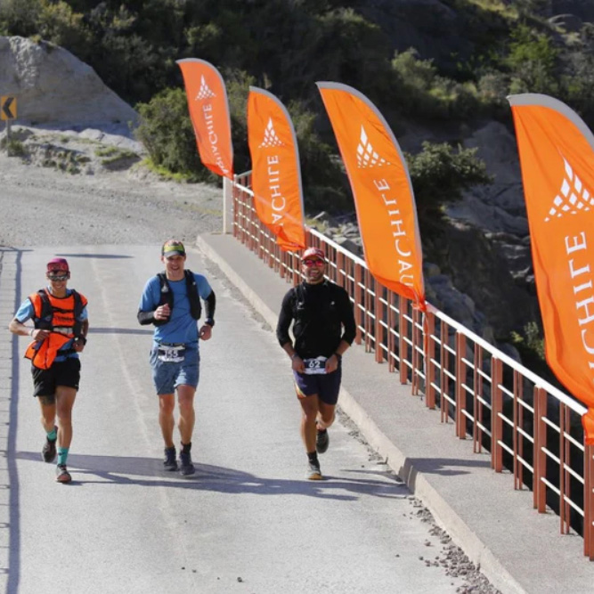 Bandera tipo pluma - Landscape image of a feather flag at a busy trade fair, pole, and base visible, crowd softly blurred, emphasizing high-visibility placement and strong print contrast for event marketing.