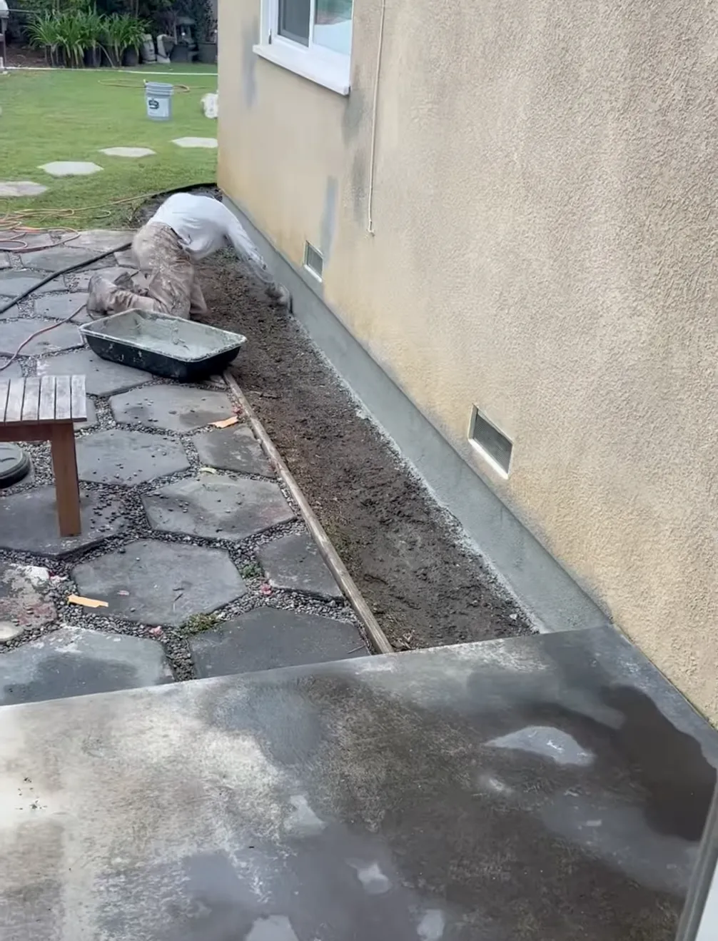 Man with grey shirt repairing grey stucco to the base of a house. 