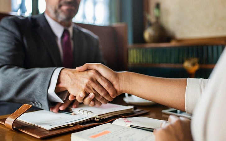 Dos personas se dan la mano sobre un escritorio en una oficina, representando una reunión profesional, acuerdo o asesoría de confianza.