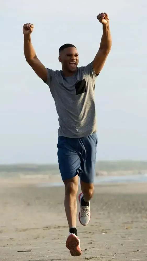 Joyful man outdoors with arms raised in celebration, wearing a grey t-shirt and blue shorts, smiling while standing on a beach or coastal area.