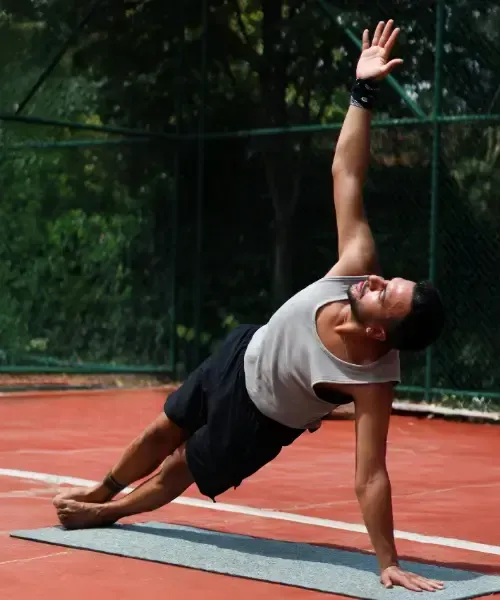 Person doing a side plank on a yoga mat on an outdoor court, one arm supporting their body and the other arm raised overhead.