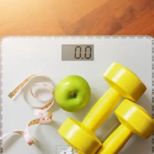 Bathroom scale on a wooden floor with a measuring tape, a green apple, and two yellow dumbbells on top.