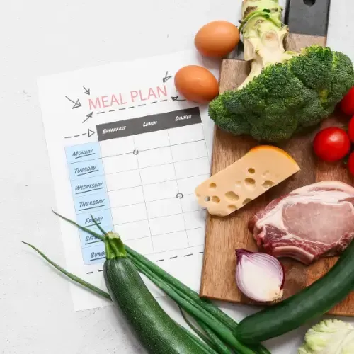 Weekly meal plan sheet next to a cutting board with eggs, broccoli, avocado, cheese, meat, and fresh vegetables.