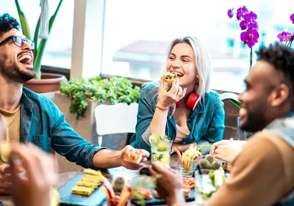 Three people laughing and eating together at a table with food and drinks.