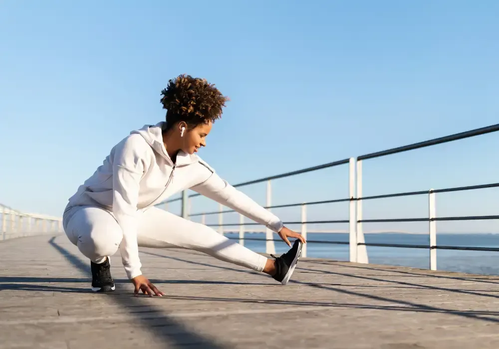 Woman in athletic clothing stretching her leg on a waterfront walkway.