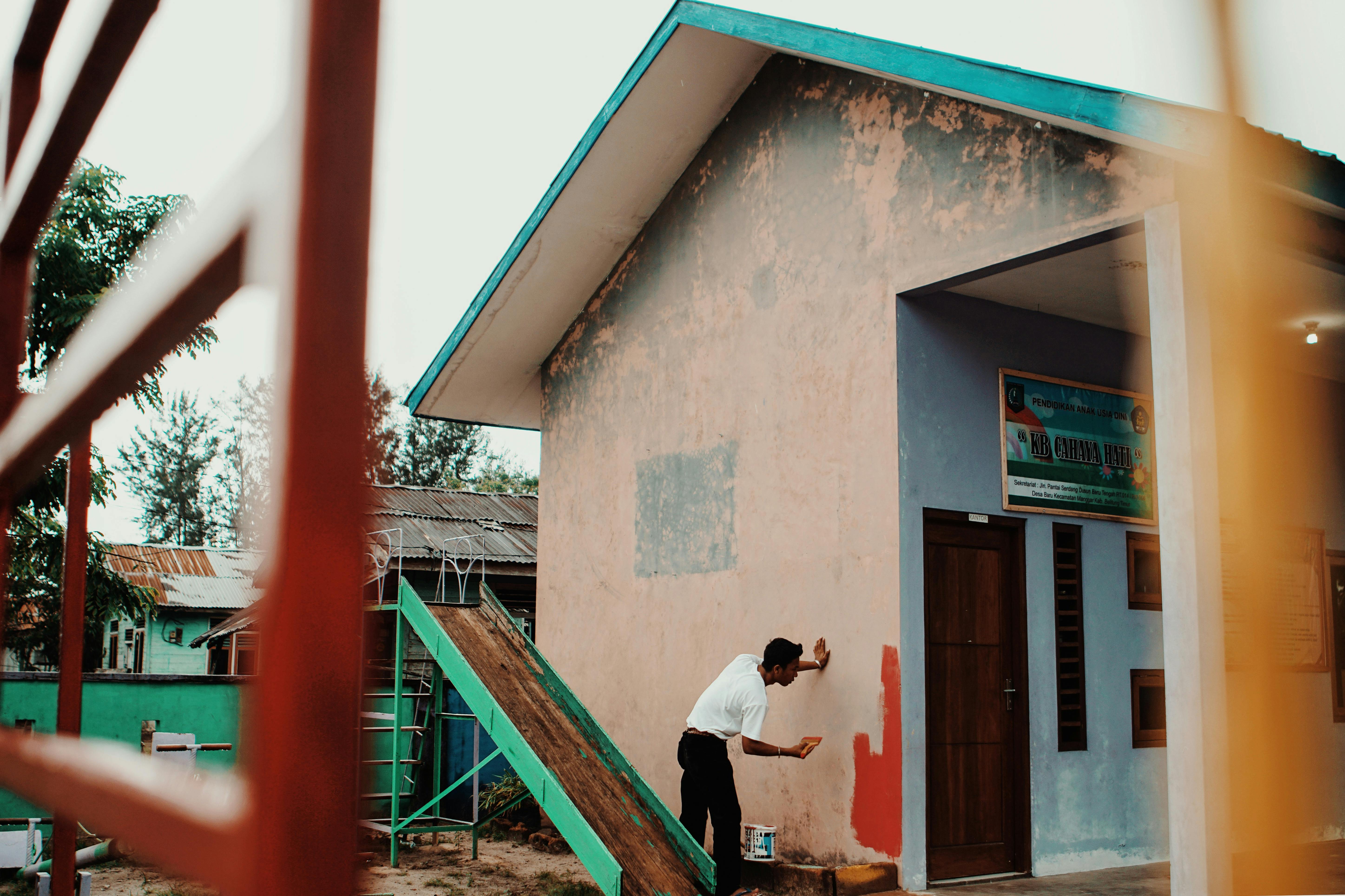white shirt man painting wall of house red using paint brush
