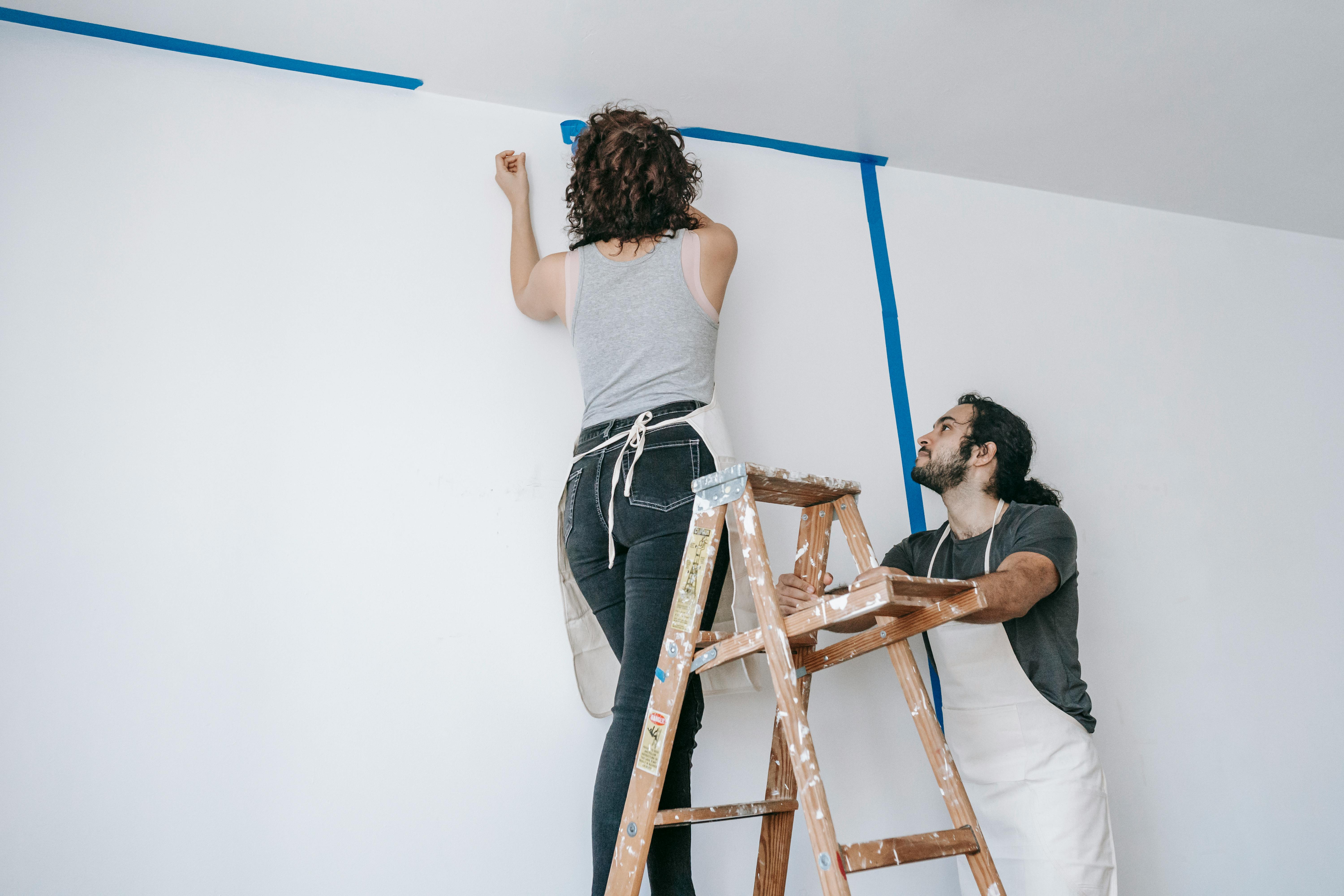 girl painting over blue tape whilst guy holds ladder