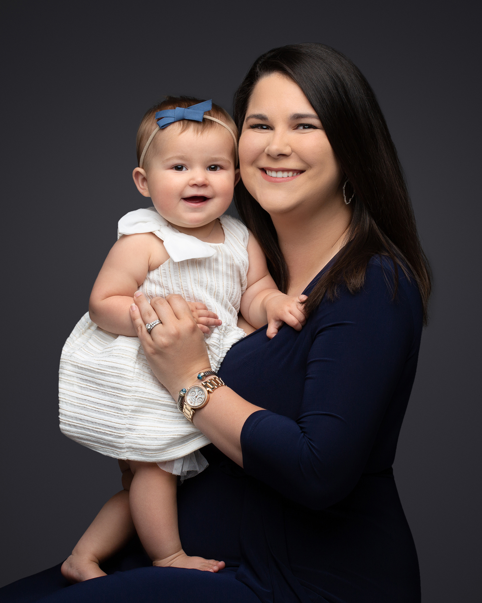 A smiling mother in a navy blue dress holding her infant daughter in a white dress for a studio portrait.