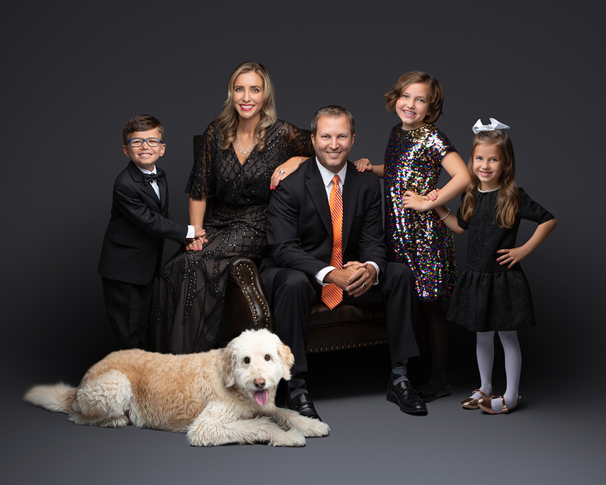 Formal family studio portrait of a father in a black suit with orange tie, mother in a beaded black gown, and three children in formal attire. A large cream-colored Goldendoodle dog lies in the foreground against a dark gray backdrop.
