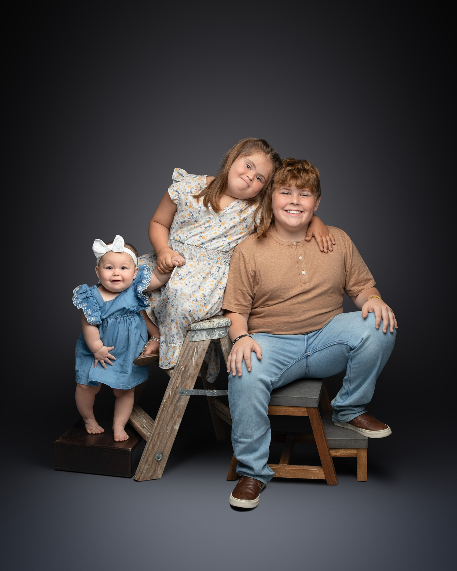 Three siblings—a baby, a young girl, and a boy—posing together on a rustic wooden ladder prop.